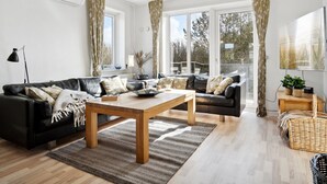 Ein modernes Wohnzimmer mit einer Eckcouch, einem Holztisch und großen Fenstern mit Vorhängen.