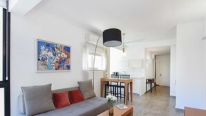 A modern living room with a grey sofa, a wooden coffee table, and a dining area with a wooden table and chairs. There is a painting on the wall and a ceiling light fixture. - Hilton Area 2BR by HolyGuest (Tel Aviv-Yafo)