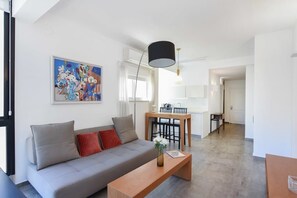 A modern living room with a grey sofa, a wooden coffee table, and a dining area with a wooden table and chairs. There is a painting on the wall and a ceiling light fixture. - Hilton Area 2BR by HolyGuest (Tel Aviv-Yafo)
