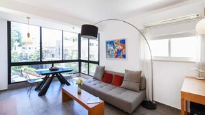 A modern living room with a sofa, a dining table, and a large window with a view of buildings outside. - Hilton Area 2BR by HolyGuest (Tel Aviv-Yafo)