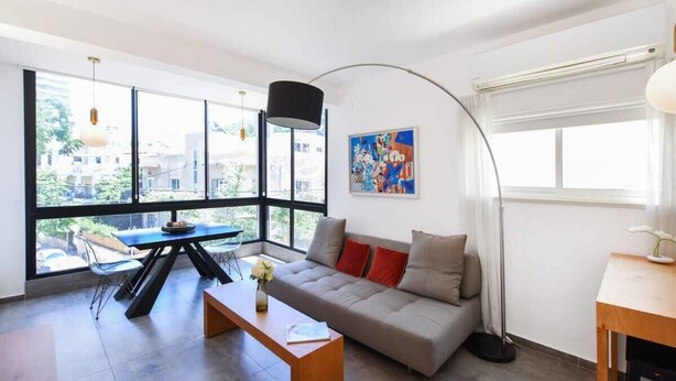 A modern living room with a sofa, a dining table, and a large window providing a view of the city. - Hilton Area 2BR by HolyGuest (Tel Aviv-Yafo)