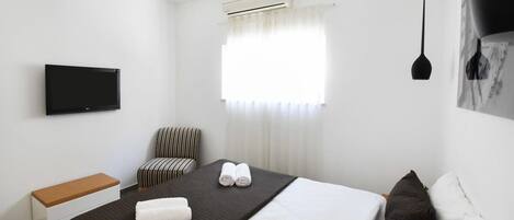 Una habitación de hotel moderna con cama, televisor de pantalla plana, banco, silla y ventana con cortinas.