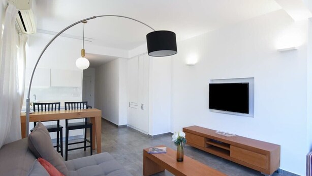 A modern living room with a sofa, a coffee table, a TV, and a dining area with chairs and a table. - Hilton Area 2BR by HolyGuest (Tel Aviv-Yafo)