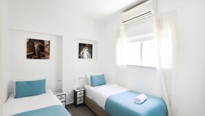 A twin room with two beds, a window with curtains, a wall-mounted air conditioner, and a small table with a lamp. - Hilton Area 2BR by HolyGuest (Tel Aviv-Yafo)