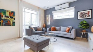 Een moderne woonkamer met een grijze bank, een grijze poef en een salontafel. Twee bijzettafels met lampen, een groot raam met gordijnen en kunstwerken aan de wanden.