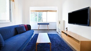 Un saló modern amb un sofà blau, una taula petita blanca, un televisor a la paret i una finestra amb vista a vegetació.