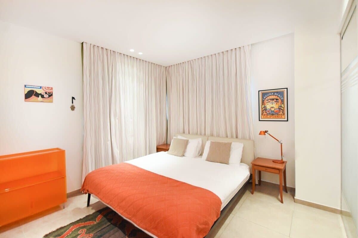 Quarto com cama, cortinas, mesa de cabeceira, luminária, quadro e um móvel laranja.