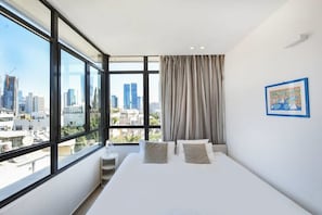 A modern hotel room with a large window offering a city view, a neatly made bed, and a framed artwork on the wall.