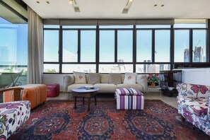 A modern living room with a large window, a sofa, a coffee table, and a patterned rug. - Spacious 2BR With Terrace in Rothschild by HolyGuest (Tel Aviv-Yafo)