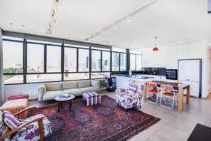 A modern living space with a sofa, ottoman, and patterned rug. A dining area with chairs and a table is adjacent to a kitchen with a refrigerator and cabinets. Large windows offer a view of the cityscape.