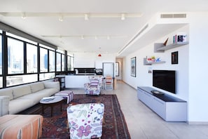 A modern living room with a sofa, ottoman, and coffee table, a kitchen area, and a large window offering a city view.