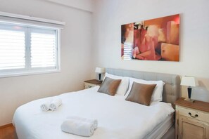 A hotel room with a bed, two bedside tables, a window with blinds, and a painting on the wall.