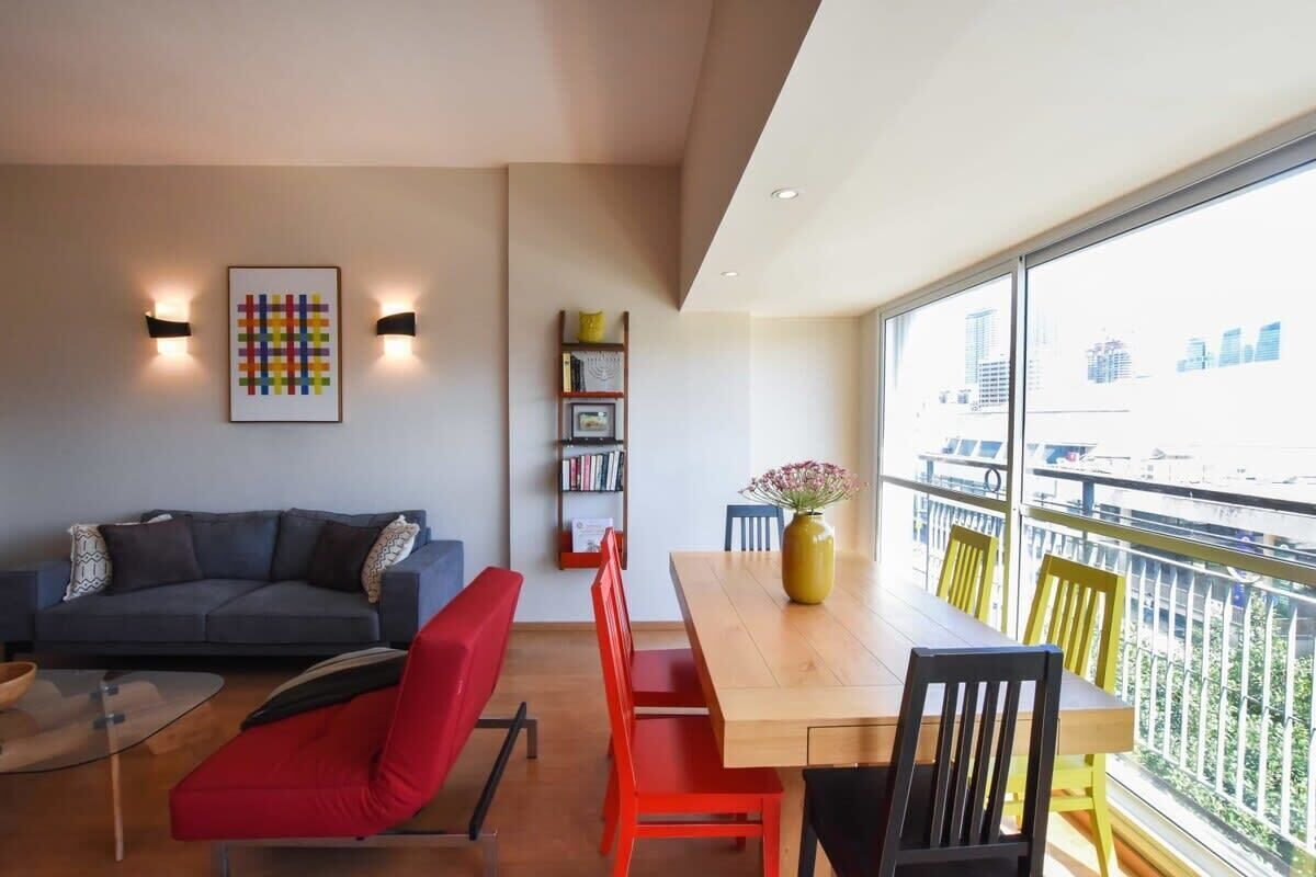 A modern living space with a sofa, red chairs, a wooden dining table, and a bookshelf.