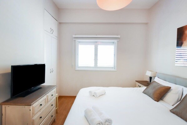 A hotel room with a bed, a TV on a dresser, a window, and a nightstand with a lamp.