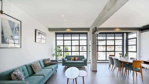 Ein geräumiges Wohnzimmer mit einem petrolfarbenen Sofa, einem runden Couchtisch und einem Esstisch mit Stühlen. Große Fenster sorgen für natürliches Licht und bieten einen Blick auf die Stadt.