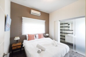 A modern hotel room with a large bed, a bedside table with a lamp, a window with blinds, an air conditioning unit, and a bathroom visible through a glass door.