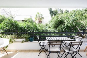 Een terras buiten met een houten tafel en stoelen, omgeven door weelderige begroeiing en een uitzicht op gebouwen op de achtergrond.