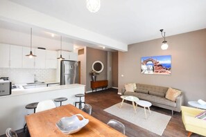 Une cuisine moderne avec des armoires blanches, un réfrigérateur en acier inoxydable et des luminaires suspendus. On y trouve un coin repas avec une table et des chaises en bois, ainsi qu’un salon avec un canapé et un tableau représentant un paysage côtier.