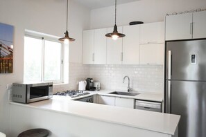 A modern kitchen with white cabinetry, stainless steel appliances, pendant lighting, and a large window providing natural light.