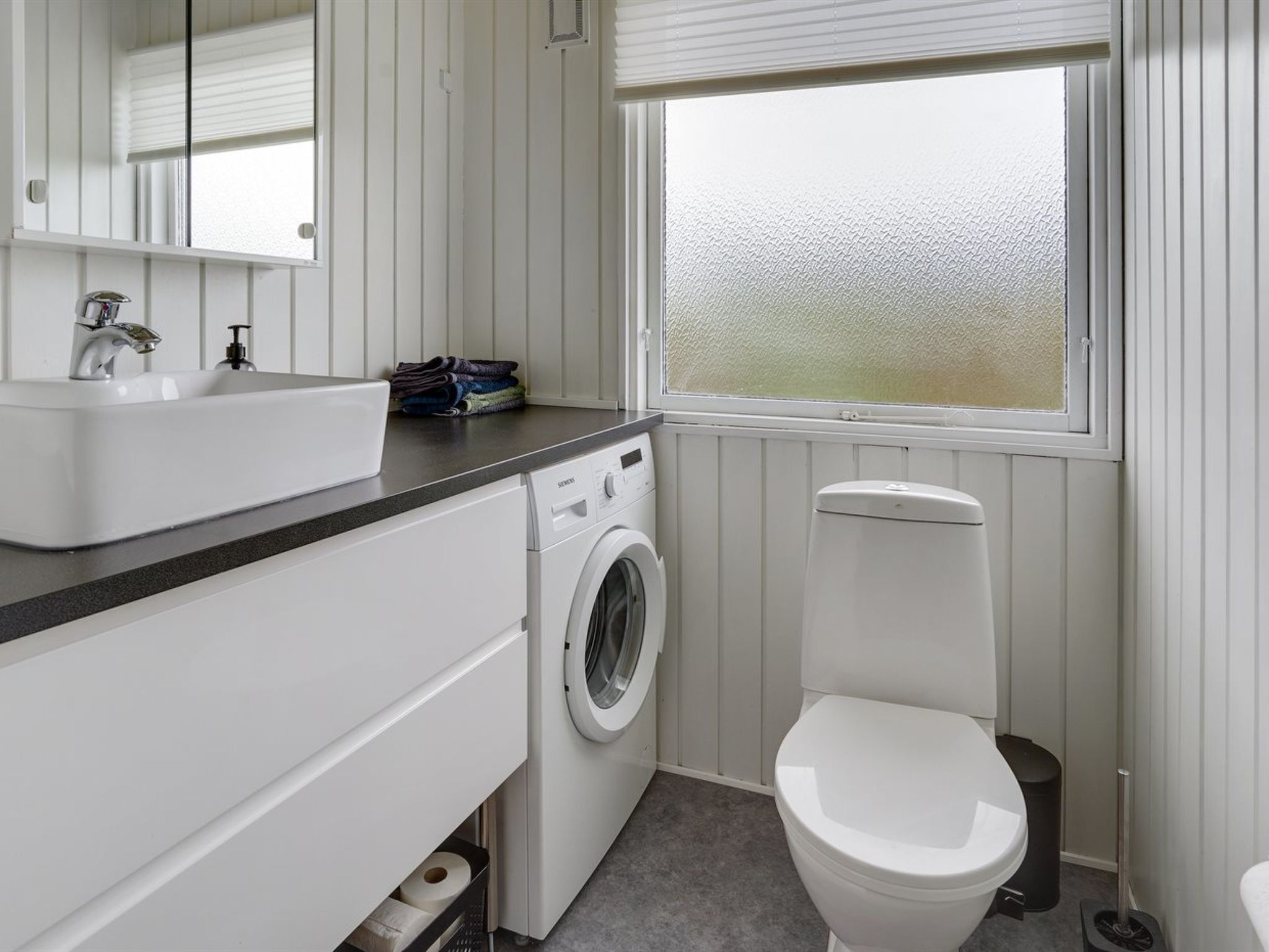 Un baño moderno y funcional con lavamanos blanco, lavadora, inodoro y ventana con vidrio esmerilado.