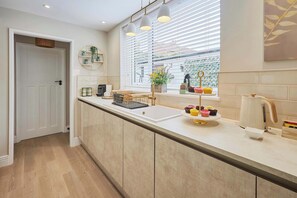 A modern kitchen with a white countertop, a sink, and a window with blinds. There are three pendant lights above the counter, and a door leading to another room. - Host & Stay | South Riggs (Bedlington)