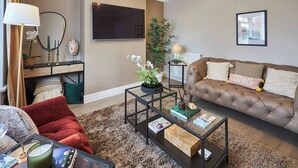 Un salon avec un canapé marron, une table basse en verre, un fauteuil rouge, un miroir, un téléviseur à écran plat, une plante en pot et un tableau encadré accroché au mur.
