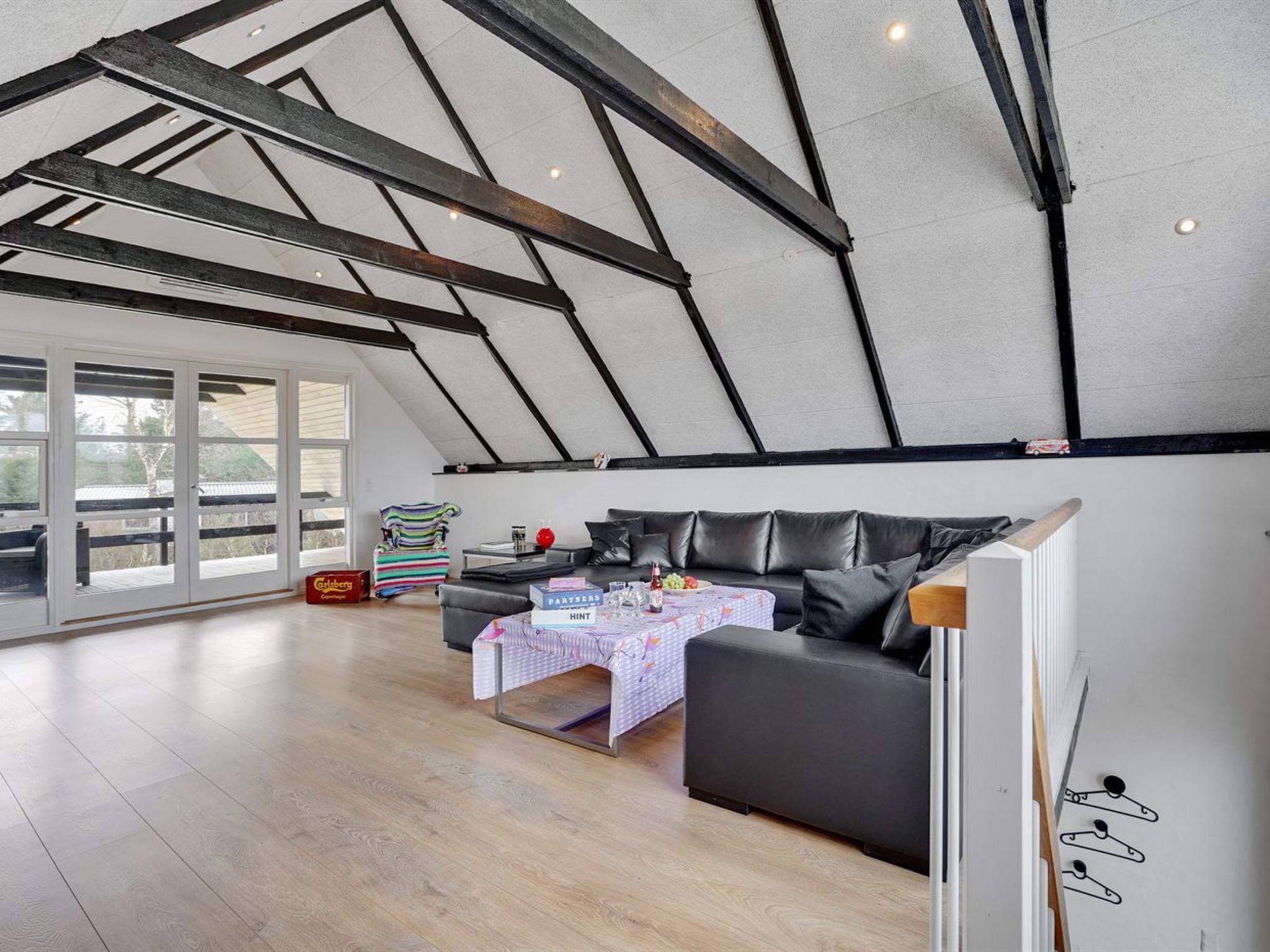 Une chambre moderne de style loft, avec un plafond en pente, un canapé en cuir noir, une petite table recouverte d’une nappe à carreaux et une fenêtre donnant sur les arbres.