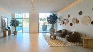 Un hall d’entrée spacieux avec une grande fenêtre, deux plantes en pot, un coin salon avec un canapé et des fauteuils, et un mur décoré de motifs circulaires.