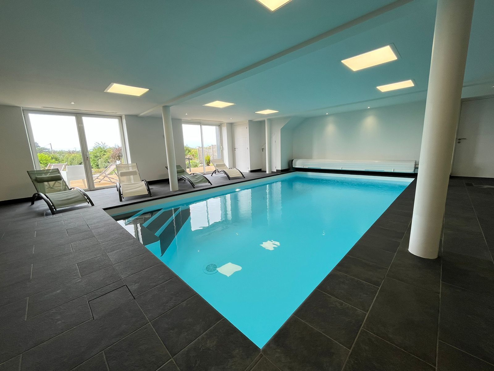 An indoor swimming pool with clear blue water, surrounded by dark tiled flooring and white walls. There are lounge chairs along the poolside, and the room has large windows allowing natural light.