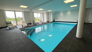 Ein Innenpool mit klarem, blauem Wasser, umgeben von dunklen Fliesenböden und weißen Wänden. Entlang des Beckens befinden sich Liegestühle, und das Zimmer verfügt über große Fenster, die natürliches Licht hereinlassen.