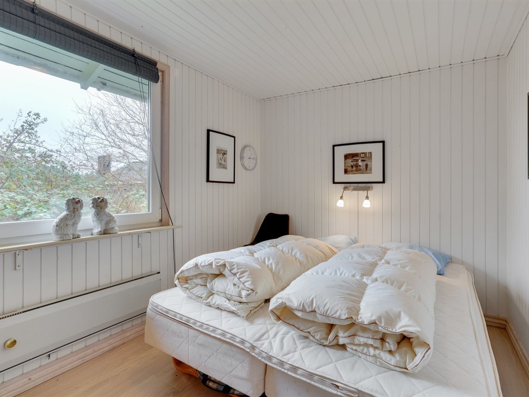 A bedroom with a bed, a window with two dogs, and two framed pictures on the wall.