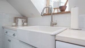 A kitchen with a white sink, a window above the sink, and a wooden shelf with kitchen items.