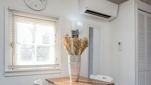 A dining area with a wooden table and white chairs, a clock on the wall, and an air conditioning unit.