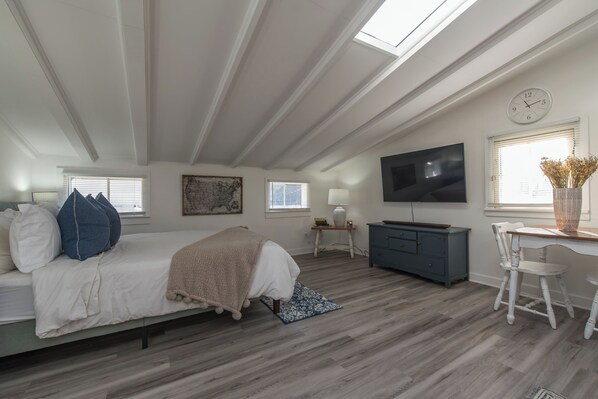 A bedroom with a bed, a desk, a TV, and a window with a clock.