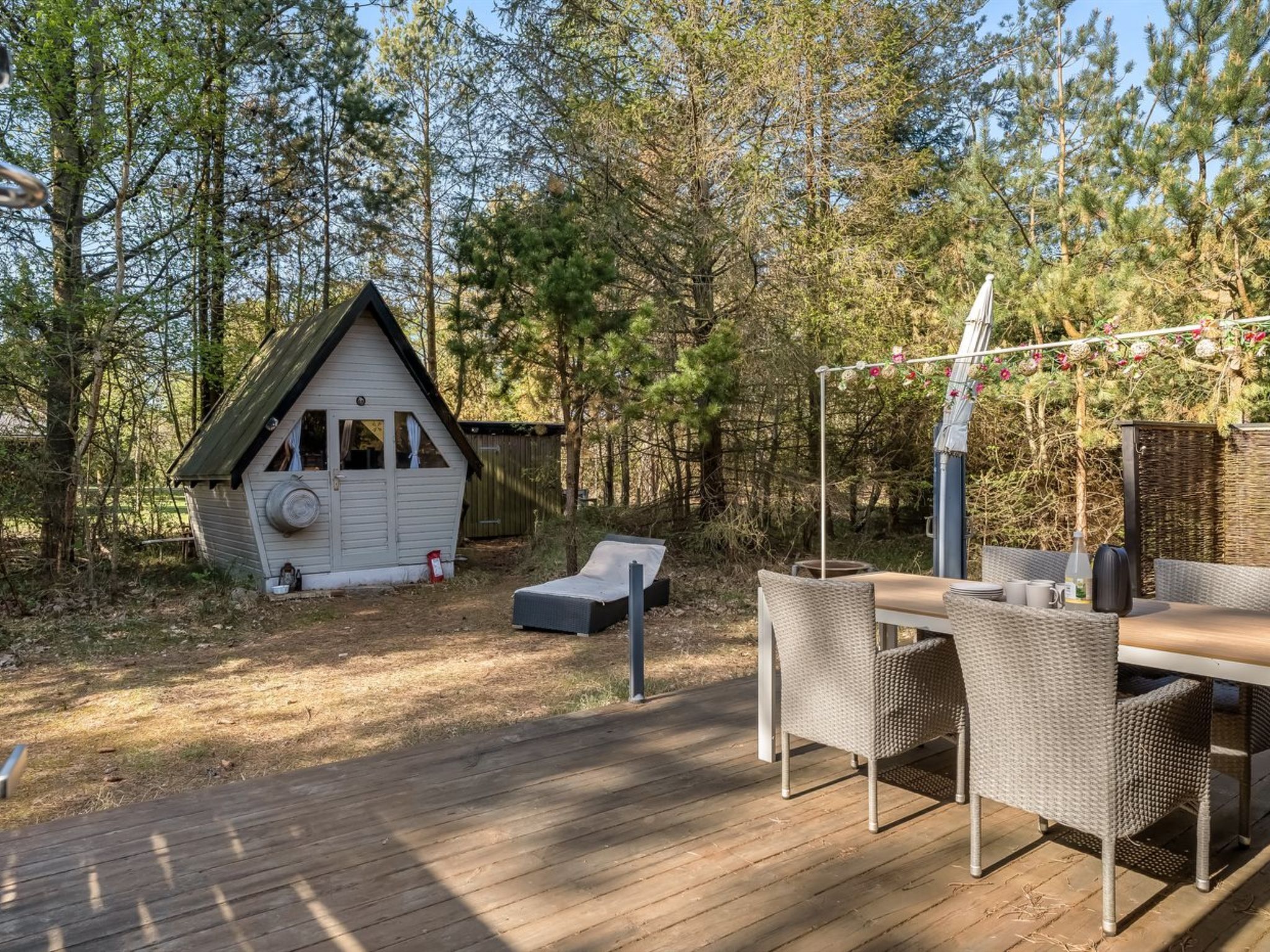 A wooden deck with a dining table and chairs, a small white cabin, and a pergola with hanging decorations.
