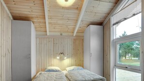 A bedroom with a wooden ceiling, two beds with white bedding, a wall-mounted light fixture, and a large window with a view of trees.