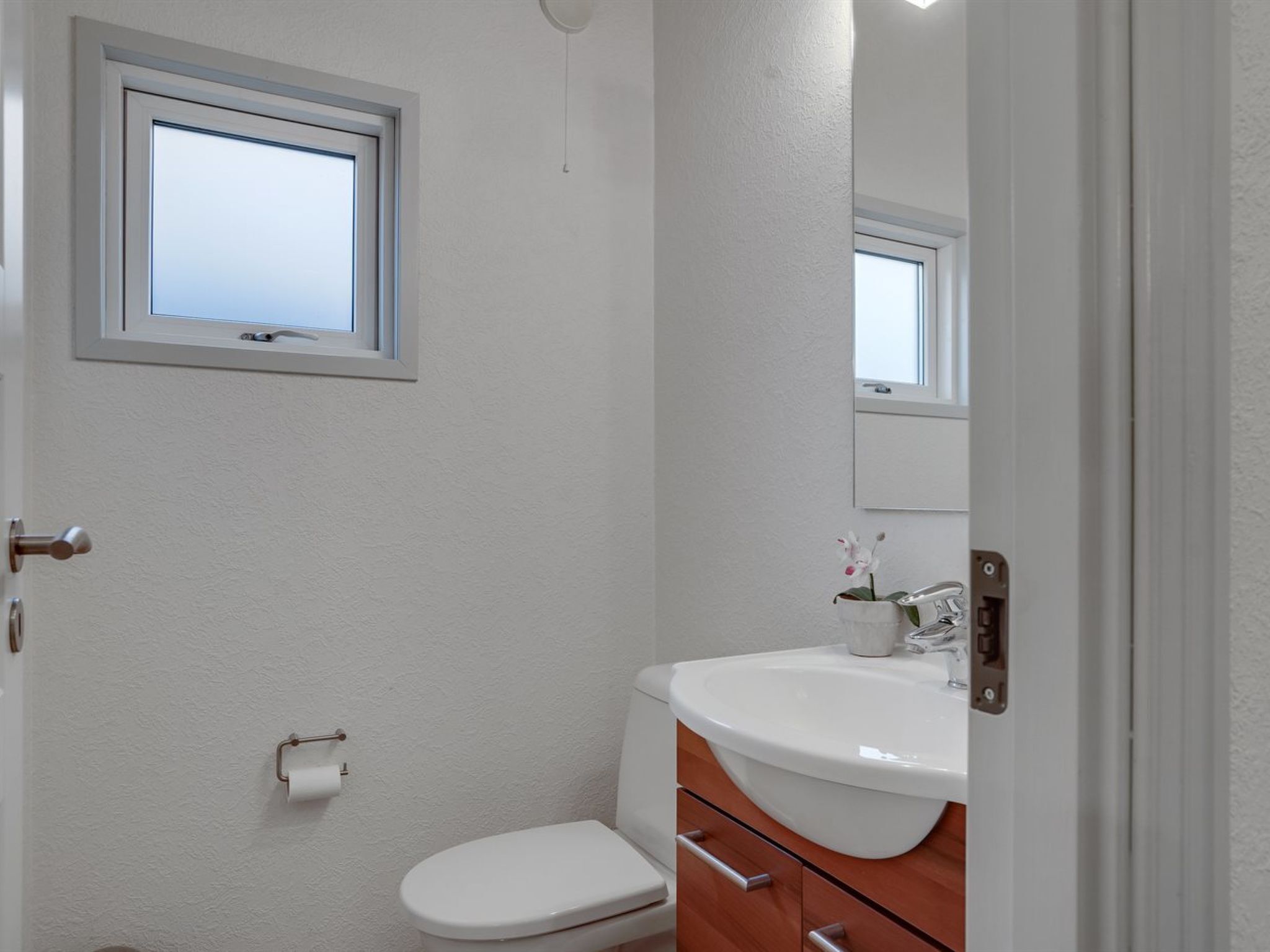 A small, white bathroom with a window, a toilet, a sink, and a mirror.