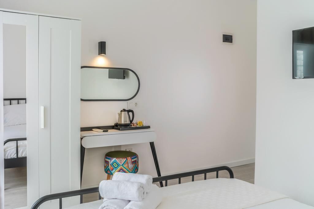 Une chambre à coucher avec un lit, une table de chevet sur laquelle se trouve une bouilloire, un tabouret coloré et un miroir accroché au mur.