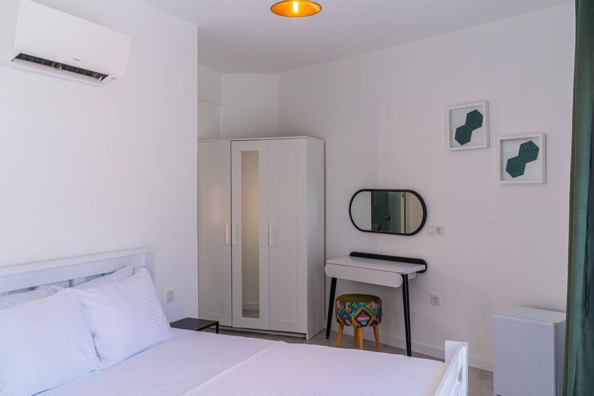 A bedroom with a bed, a wardrobe, a desk with a mirror, and a small stool.