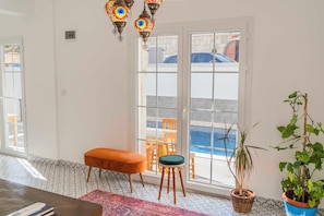 A room with a large sliding glass door leading to a pool area, a wooden bench, a small stool, and a potted plant. - Mea Reisdere (Cesme)