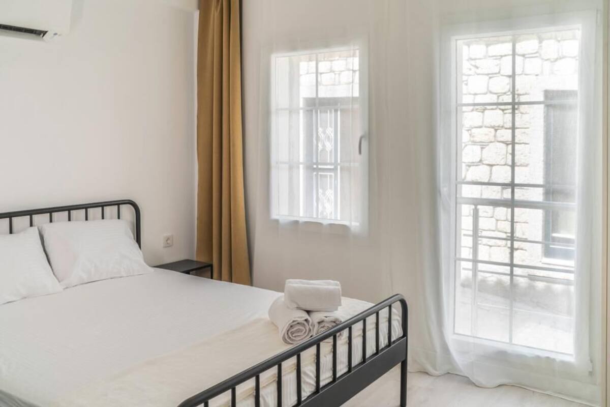 A neatly made bed with white linens and a rolled towel, a black metal headboard, a nightstand with a lamp, and a window with a view of a stone wall.