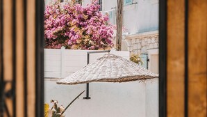 A view of a white building with a pink flowering tree and two beige lounge chairs under a woven umbrella. - Mea Reisdere (Cesme)