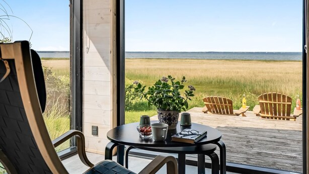 A room with a large window overlooking a beach, a round table with a book and a cup, and two wooden chairs.