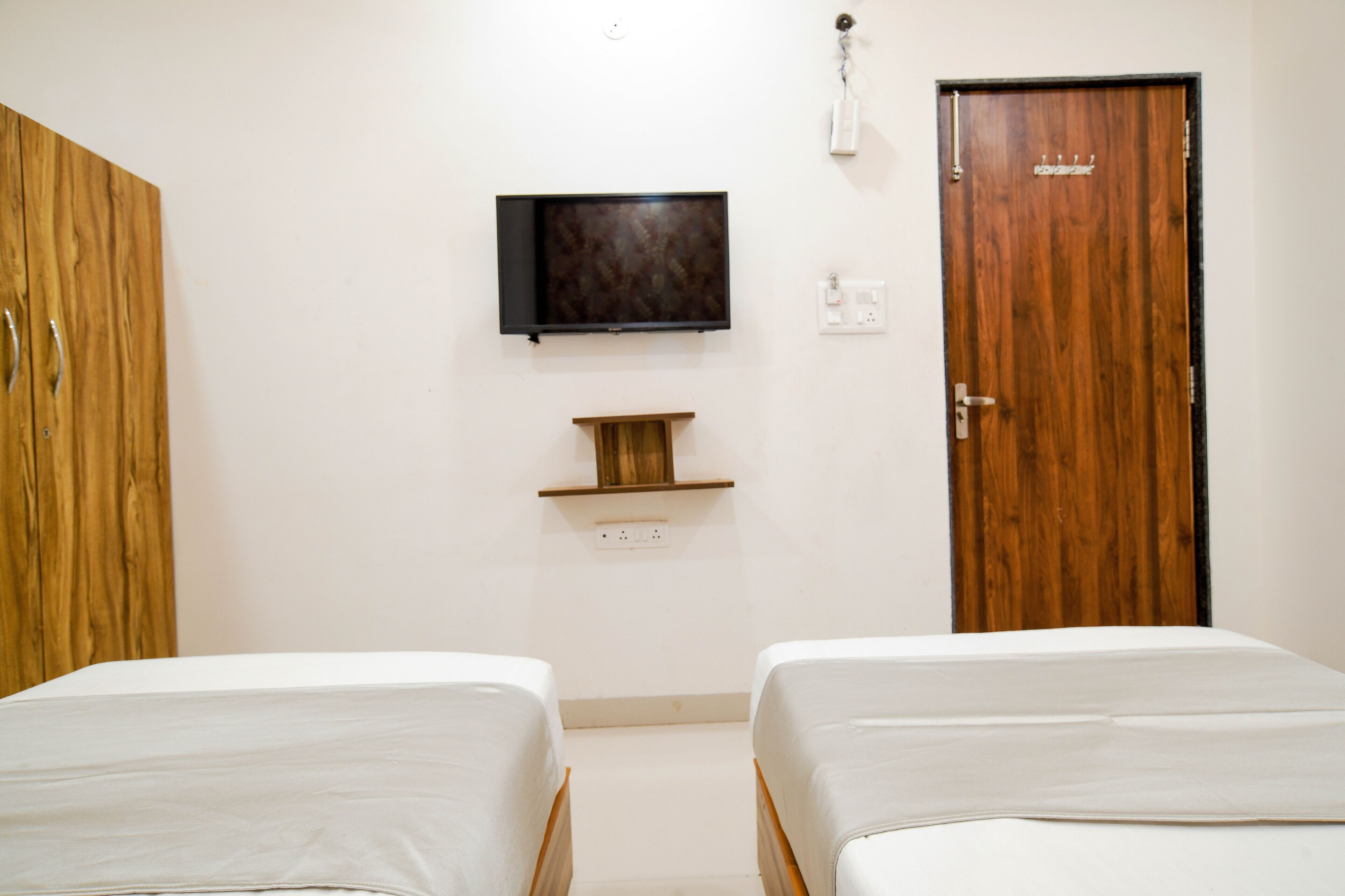 Une chambre d’hôtel avec deux lits, un téléviseur fixé au mur, une porte en bois et une petite étagère située sous le téléviseur.