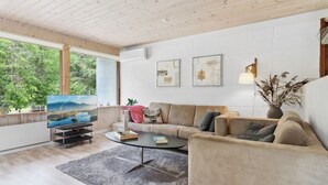 Un salon moderne avec un plafond en bois, une grande fenêtre donnant sur un espace verdoyant, un canapé beige, une table basse et un téléviseur à écran plat.