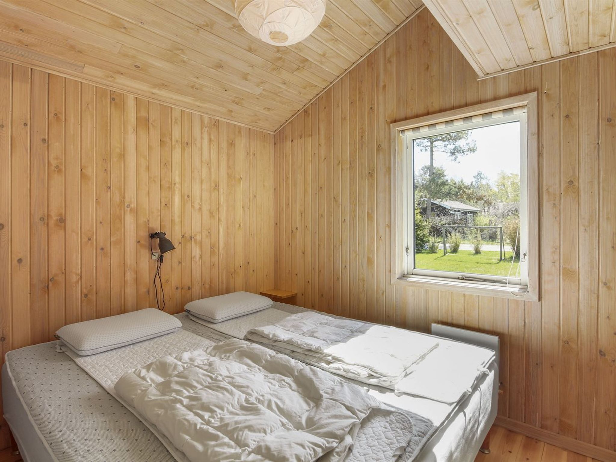 Een slaapkamer met houten lambrisering, met een enkel bed, een nachtkastje, een lamp en een raam met uitzicht op een tuin.