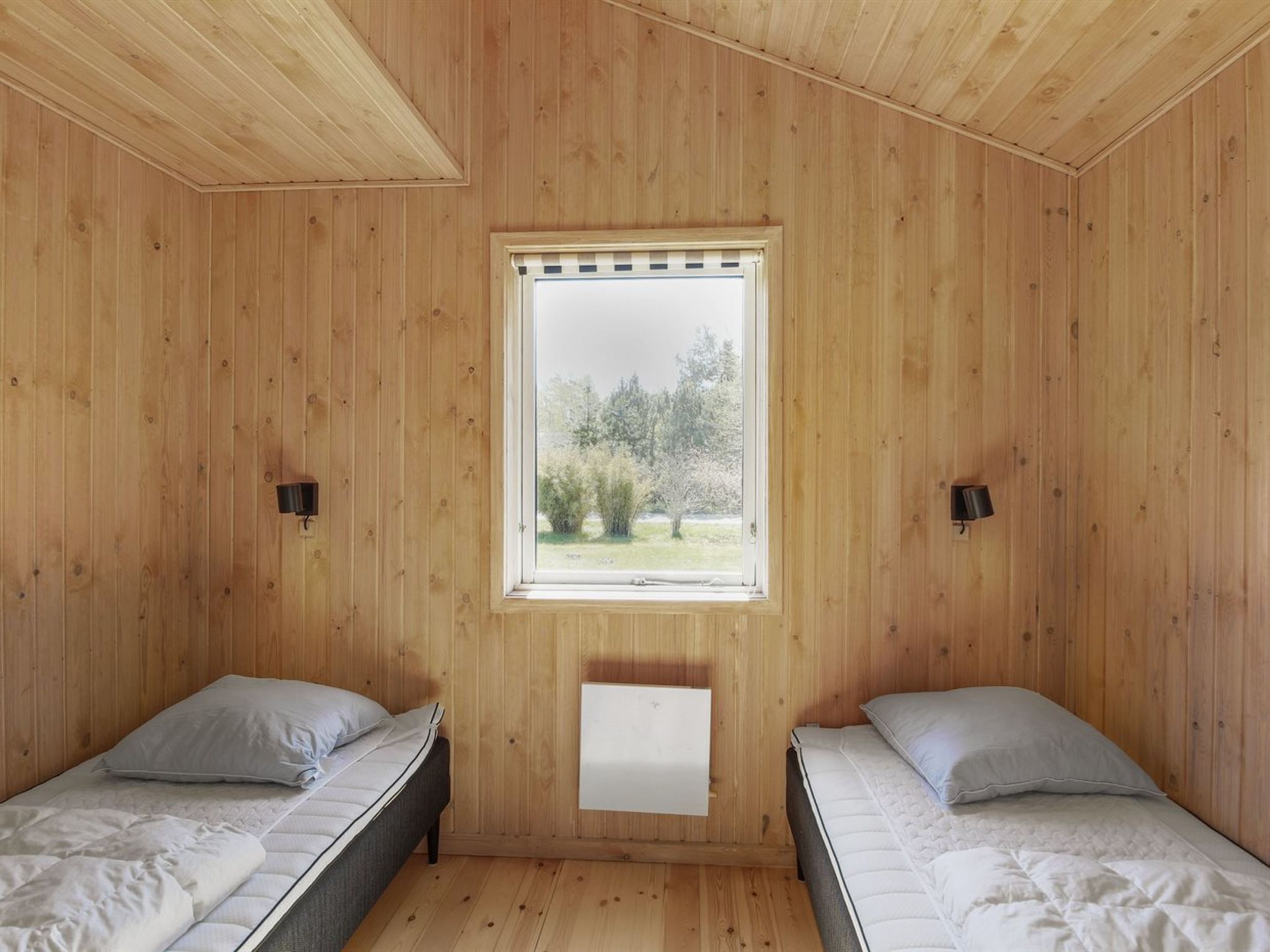 A room with two beds, wooden walls, a window with a view of trees, and wall-mounted lamps.