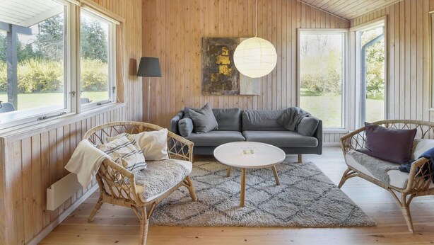 Een gezellige kamer met houten wanden, een grijze bank, rieten stoelen en een ronde tafel.