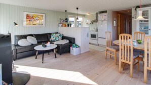 Un salon chaleureux avec un canapé, une table basse et un coin repas avec des chaises. On aperçoit une cuisine équipée au fond de la pièce.