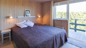 Une chambre avec un plafond en bois, un lit recouvert d’une couvre-lit à motifs sombres, une table de chevet, un tableau encadré posé sur une étagère et une fenêtre donnant sur un paysage verdoyant.